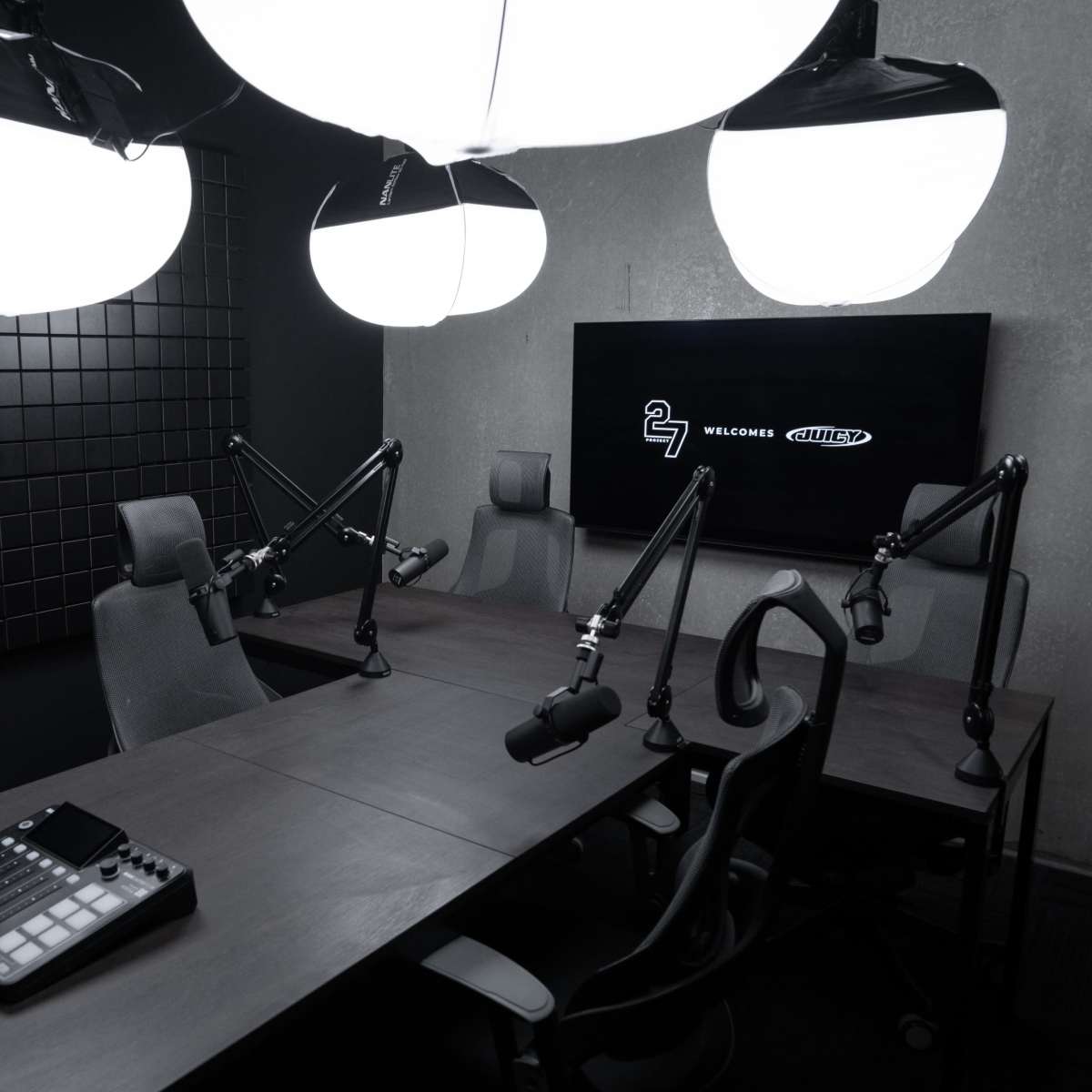 A modern podcast studio featuring a large wooden table with microphones and an audio mixer, accompanied by three adjustable chairs and dramatic overhead lighting.