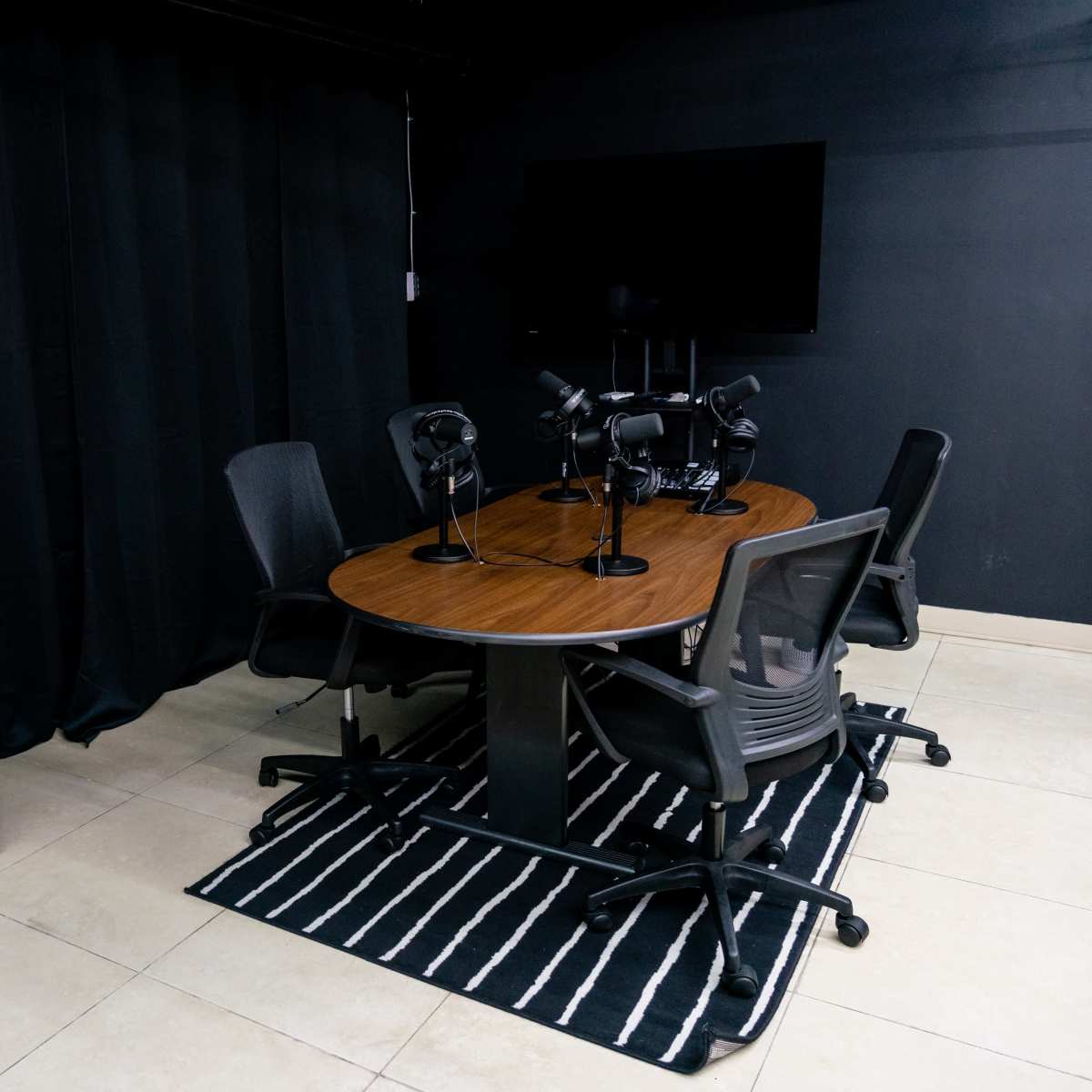 A conference room features a round table with four chairs, microphones positioned around it, and a large TV screen on the wall, set against a backdrop of black curtains.