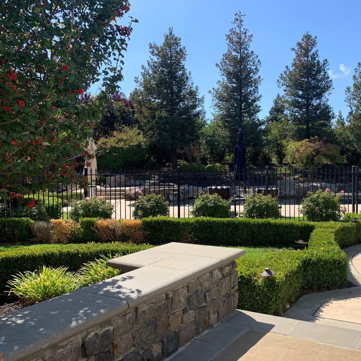 A landscaped garden with neatly trimmed hedges, stone pathways, and a fenced pool area surrounded by trees and flowering plants.