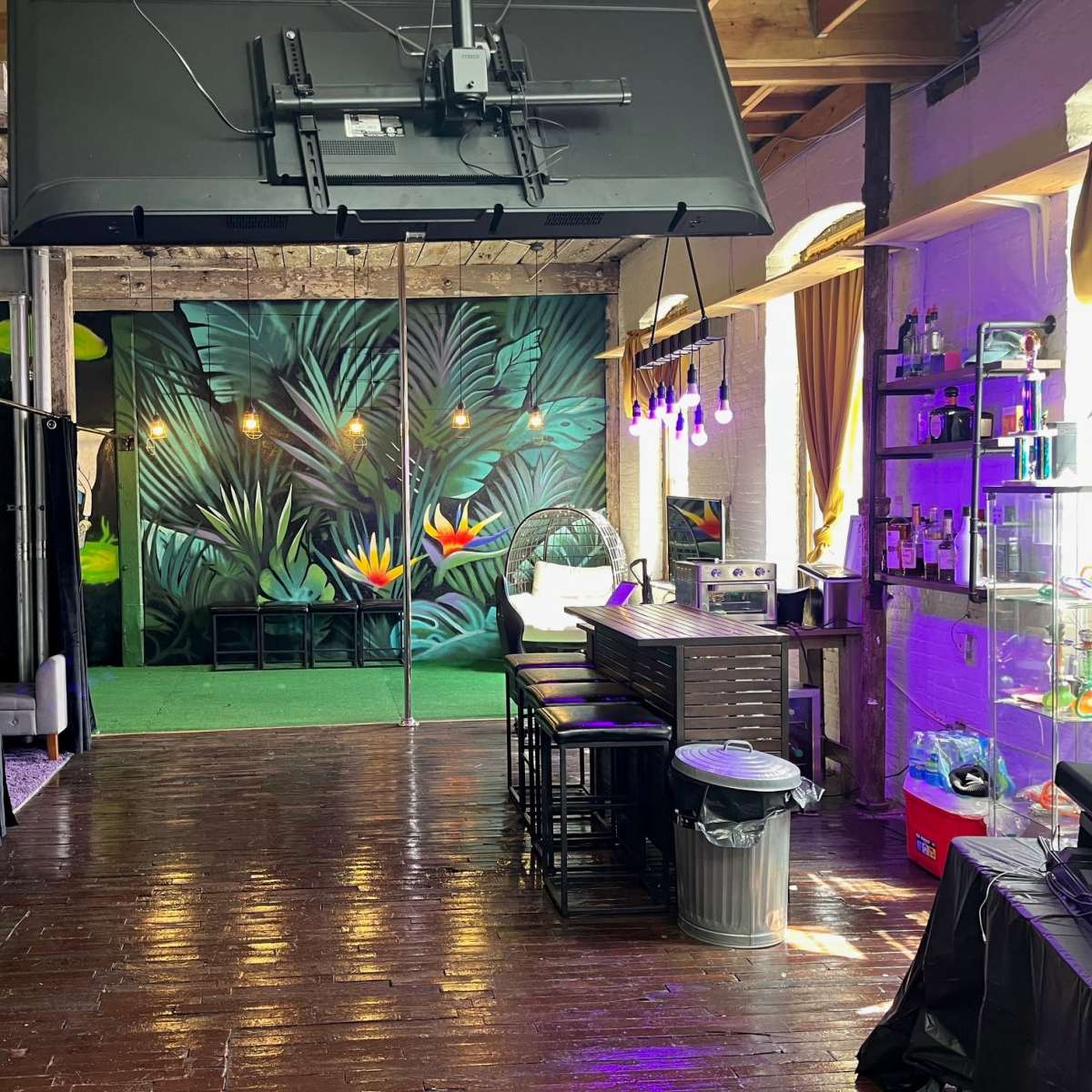 The interior of a spacious venue features a large wall mural of tropical plants, a bar area with seating, and subtle lighting.