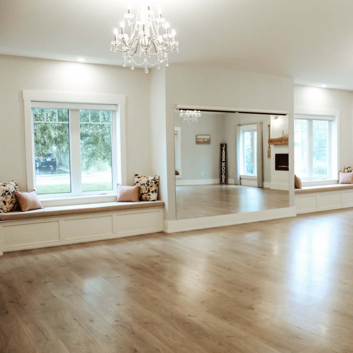 A spacious room with wooden floors, large windows, and two decorative chandeliers, featuring mirrored walls and built-in seating areas.