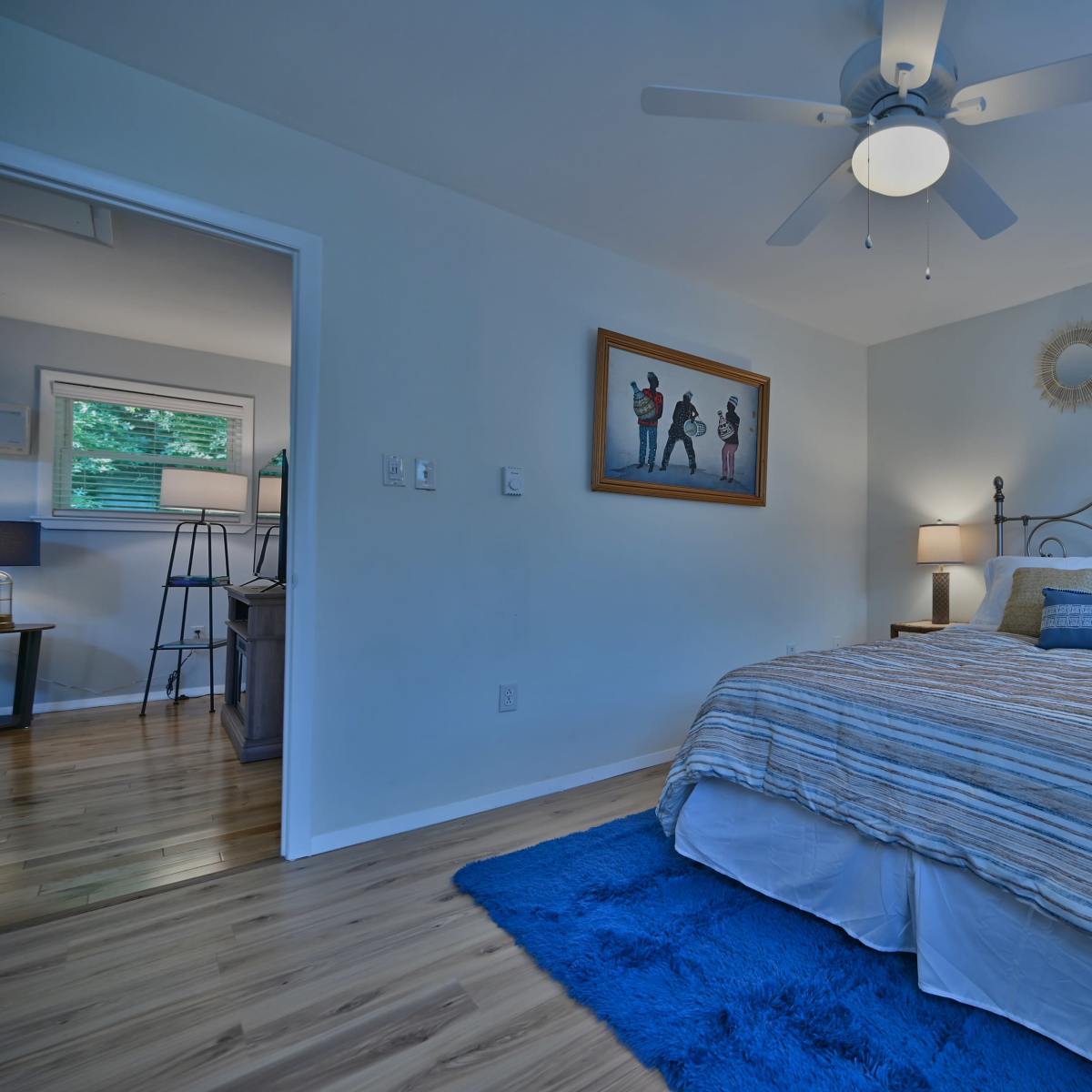A bright, minimalist bedroom with a bed covered in striped bedding, a blue area rug, and framed artwork on the wall.