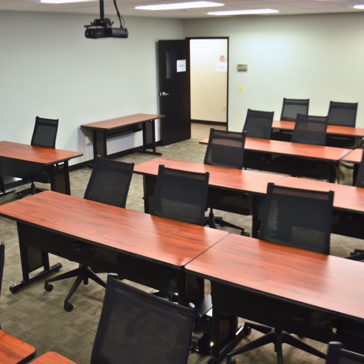 A classroom is set up with multiple rows of wooden tables and black mesh chairs arranged for a lecture or meeting.