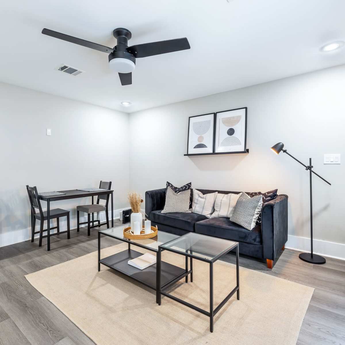 A modern living space featuring a dark sofa, a glass coffee table, a dining area with a table and chairs, a small fridge, and a floor lamp.