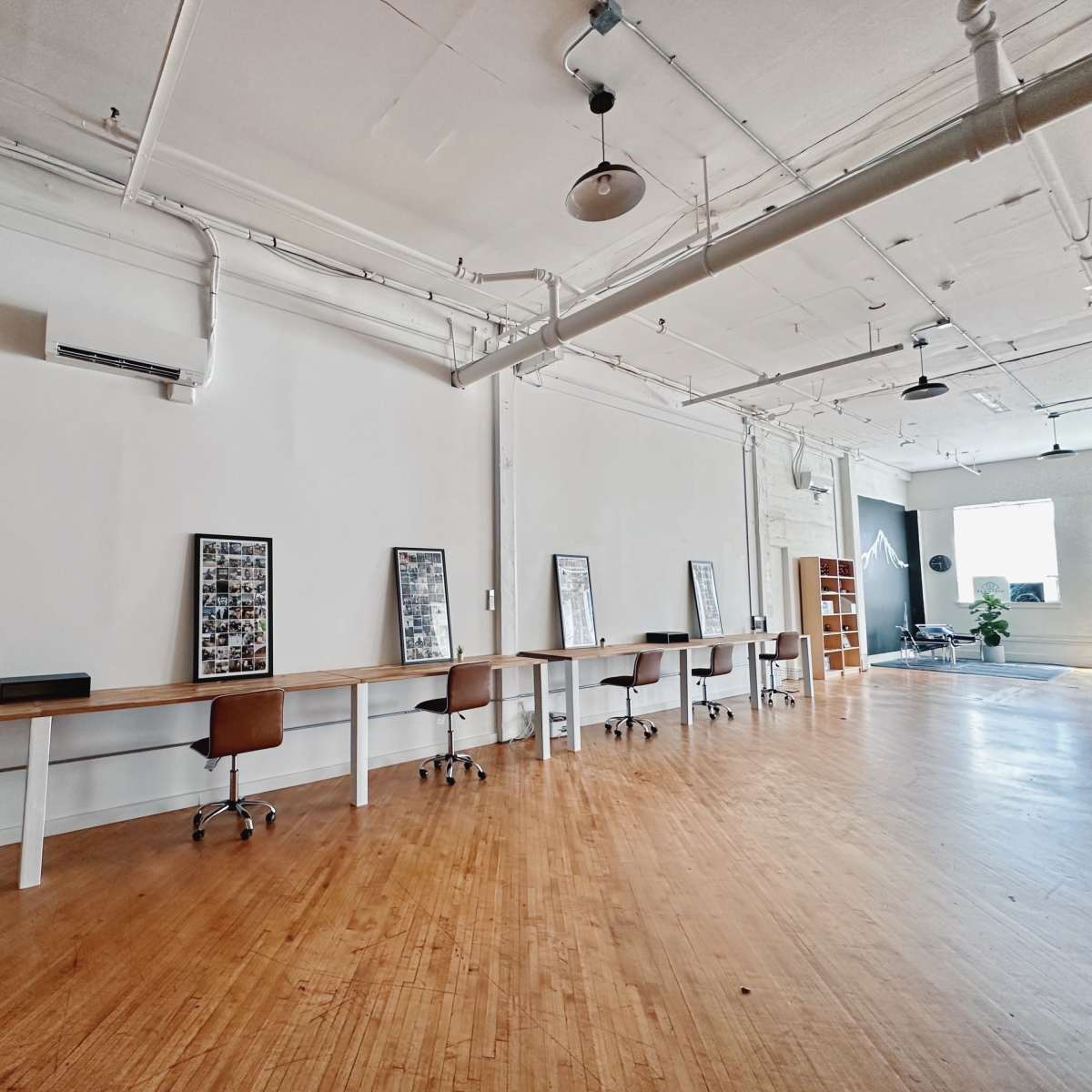 A spacious, minimalist office interior with wooden flooring, featuring several work desks and framed photos on the walls.