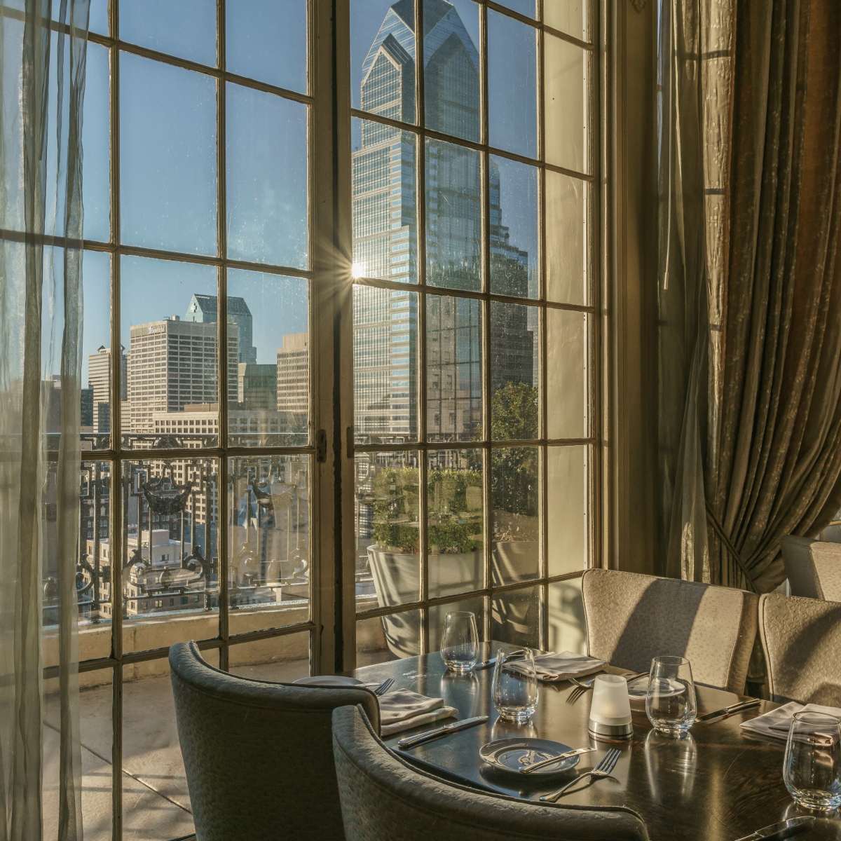 A dining area features a polished table set with glassware, overlooking a city skyline through large windows.
