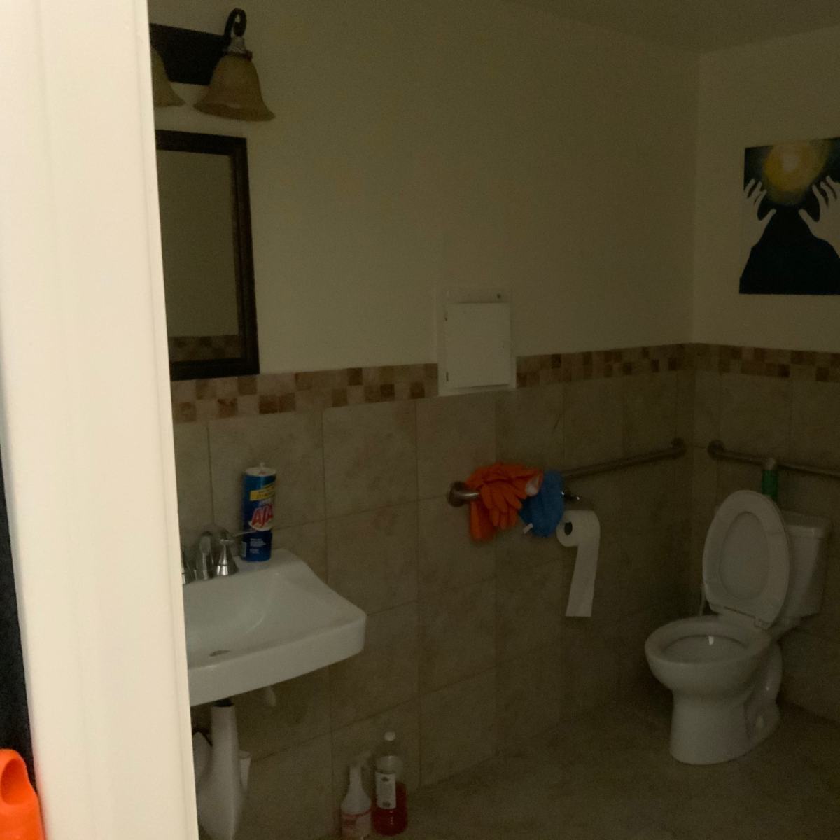 A small bathroom featuring a toilet, a sink with a mirror, and a painting on the wall, with tiled walls and a roll of toilet paper visible.