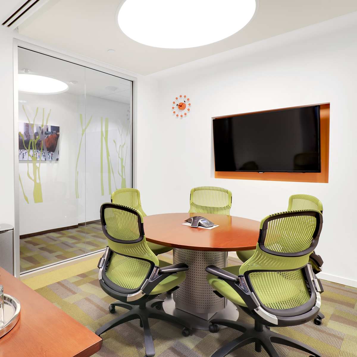 A modern conference room features a round table surrounded by green office chairs, a large wall-mounted screen, and glass walls adorned with plant graphics.