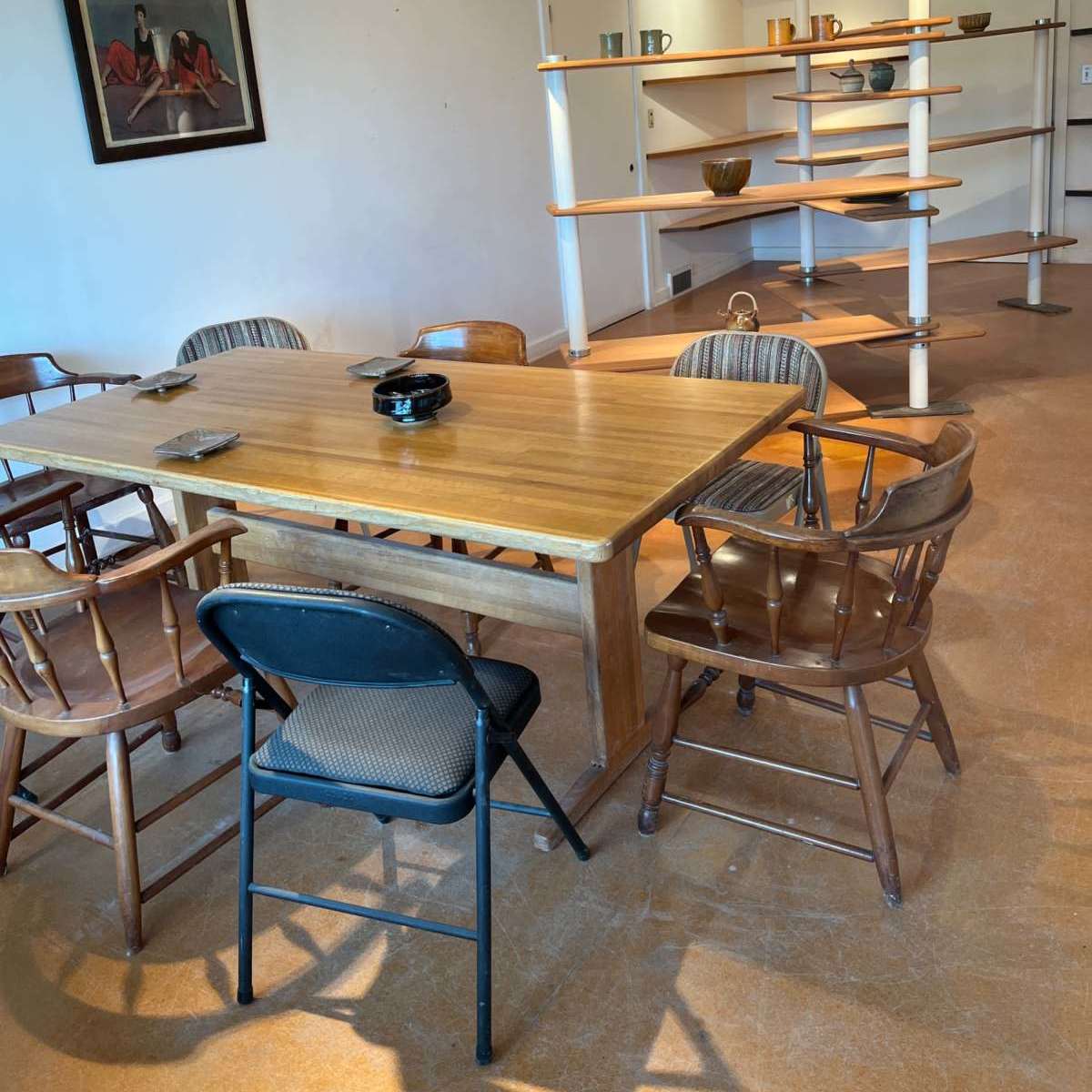 A wooden table with several chairs is placed in a brightly lit room featuring shelves displaying various items.