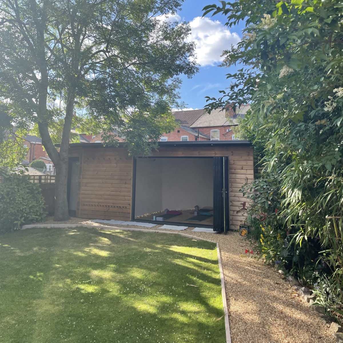 A wooden structure with sliding doors is situated in a garden surrounded by trees and shrubs under a partly cloudy sky.