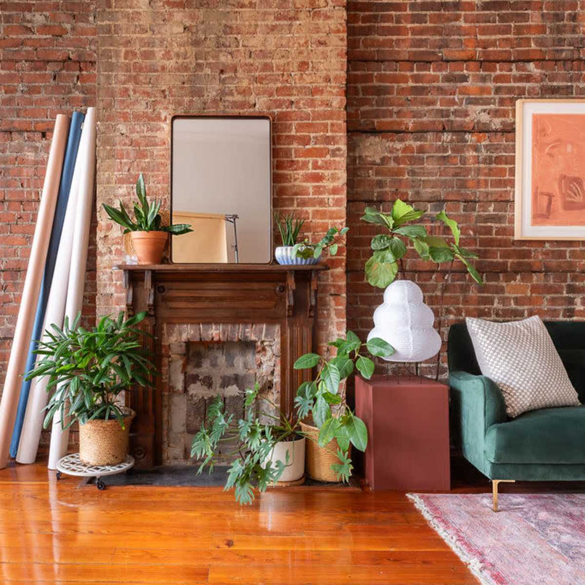 A cozy interior space featuring a brick wall, a green sofa, a desk with an office chair, plants, and a fireplace with decorative items.