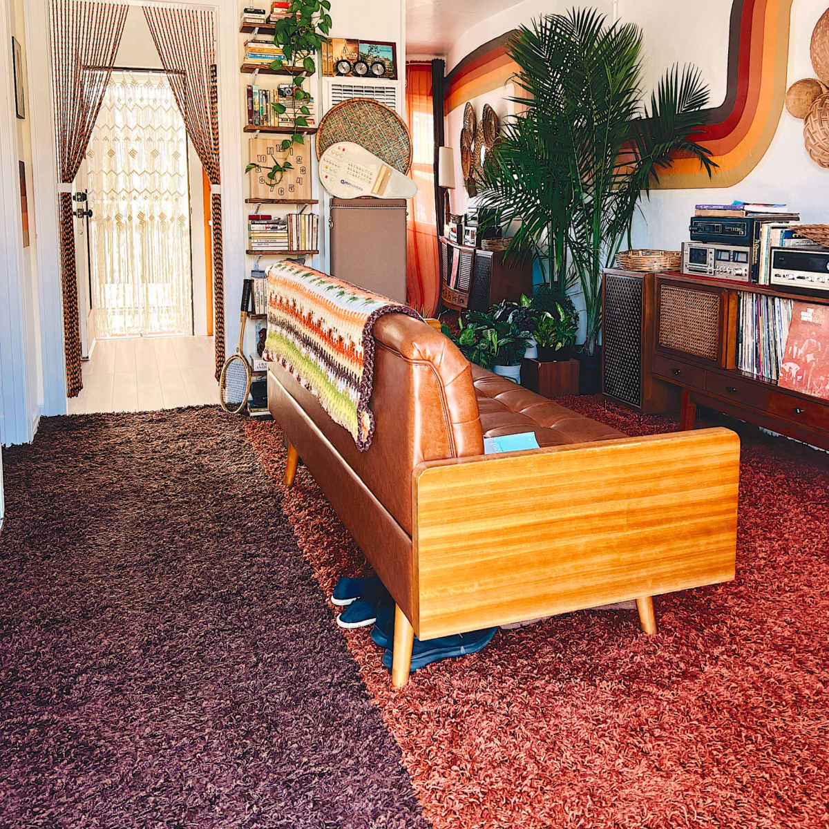 A mid-century modern living room features a wooden sofa with a colorful blanket, plants adorning the walls and shelves, and a record player setup beside a collection of vinyl records.