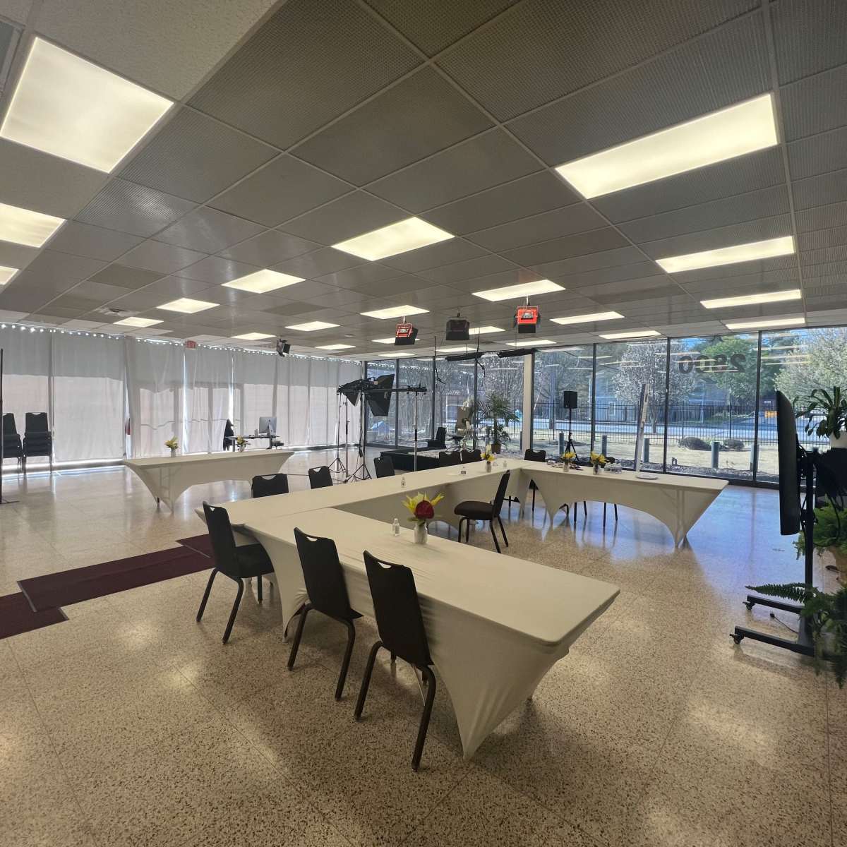 A spacious room is set up with several tables covered in white linens, surrounded by black chairs, and large windows that let in natural light.