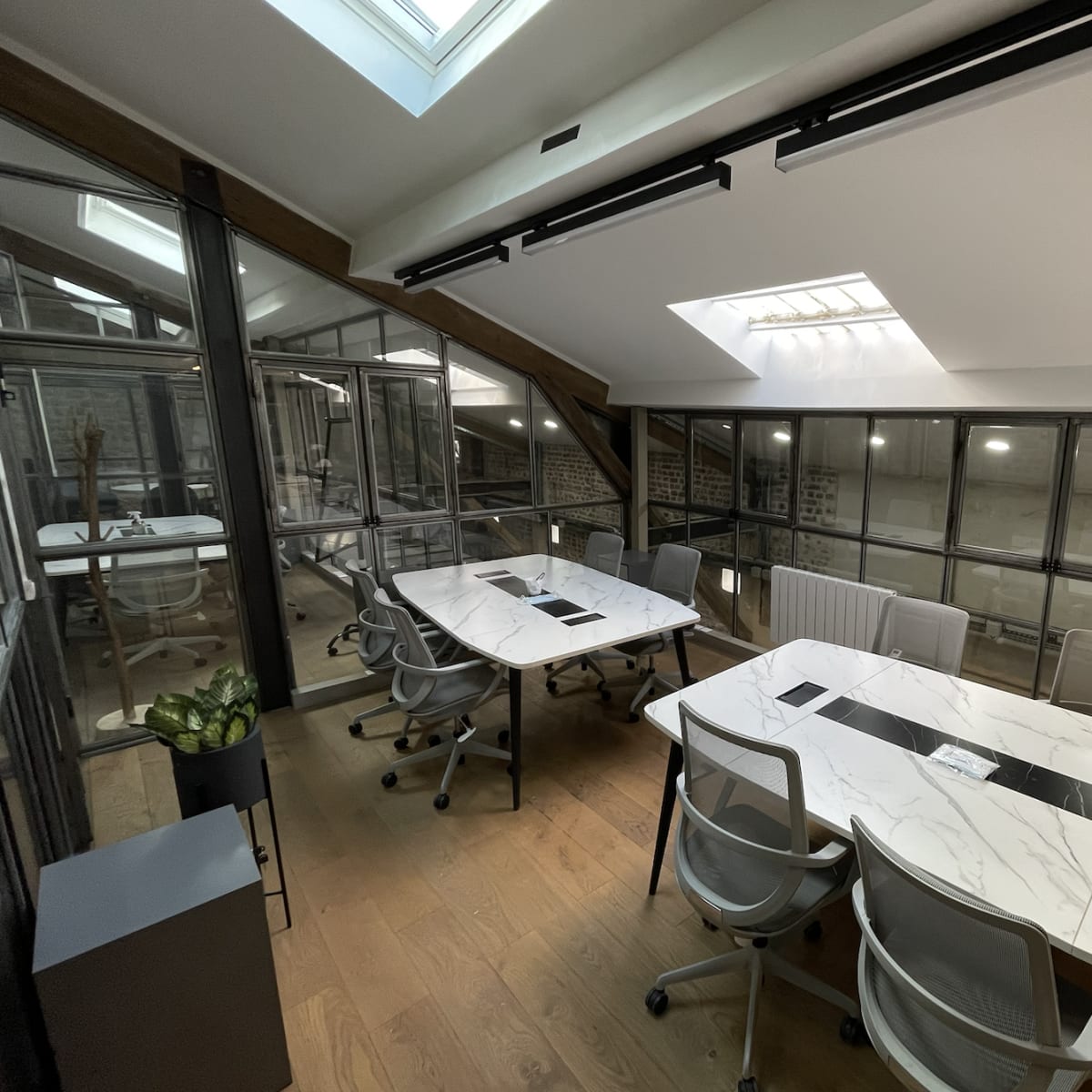 A modern office space with two white marble conference tables, surrounded by glass walls and wooden beams.
