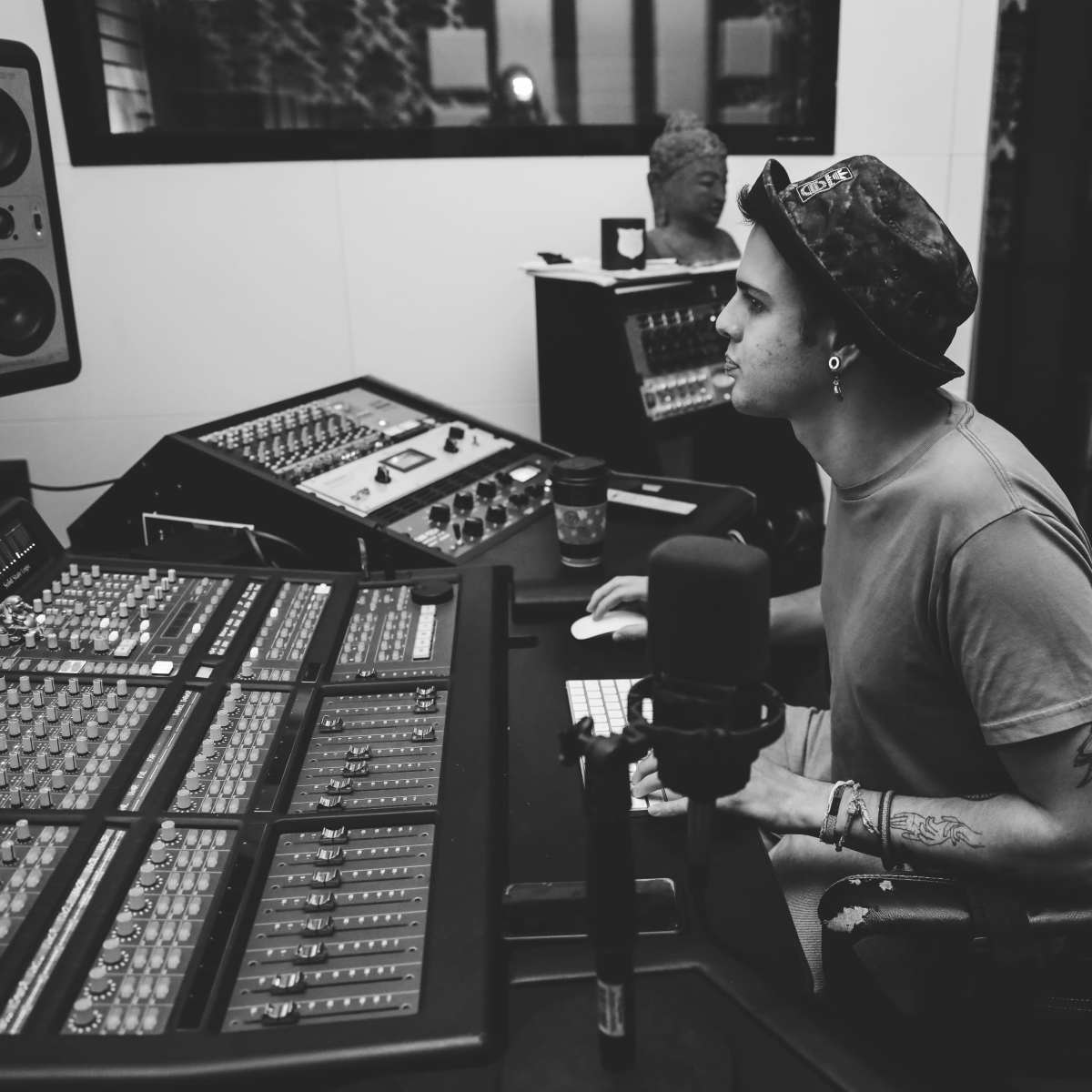A person is seated at a recording studio console, intently working on a computer while surrounded by audio equipment.