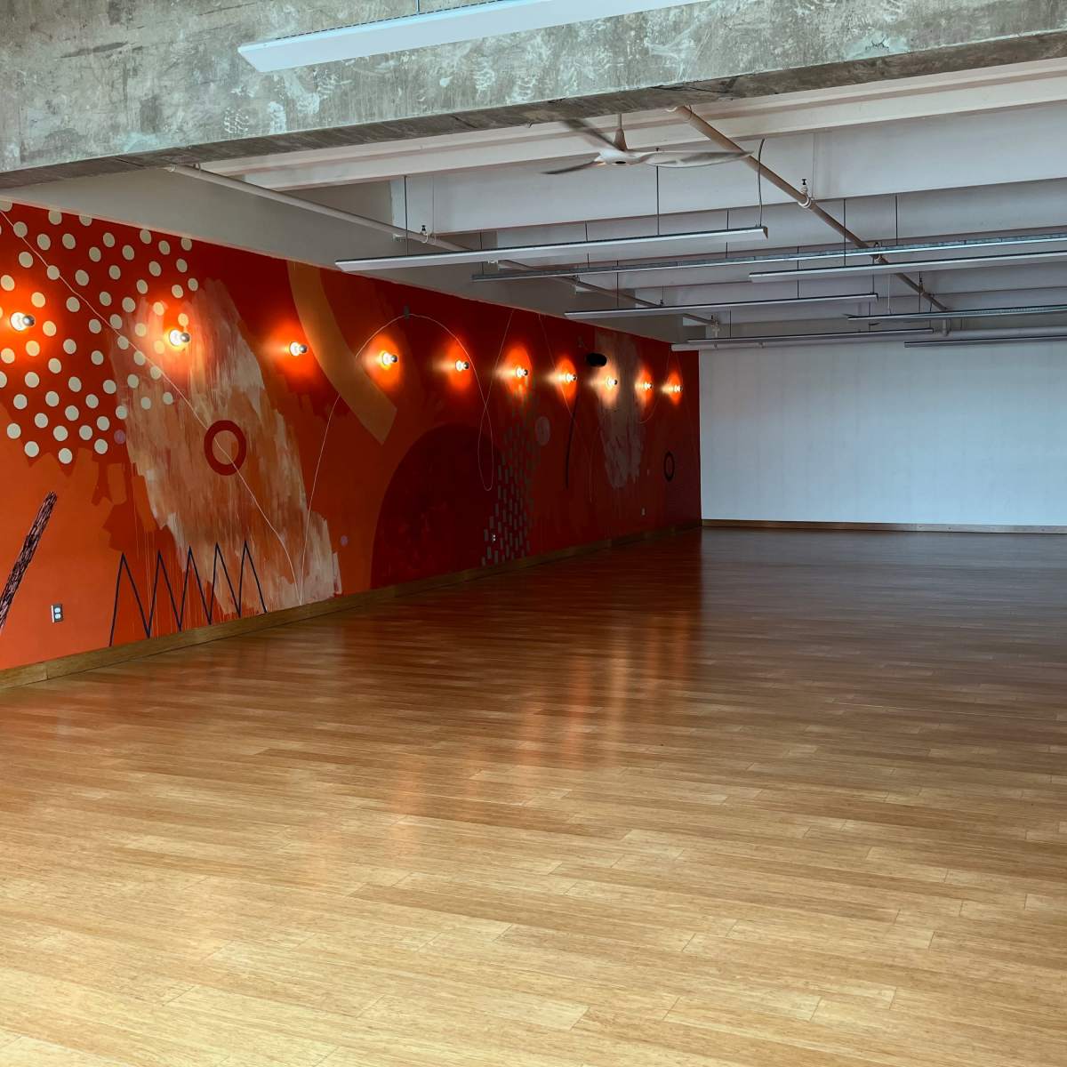 An empty room with a wooden floor, a colorful mural on one wall, and bright overhead lighting.