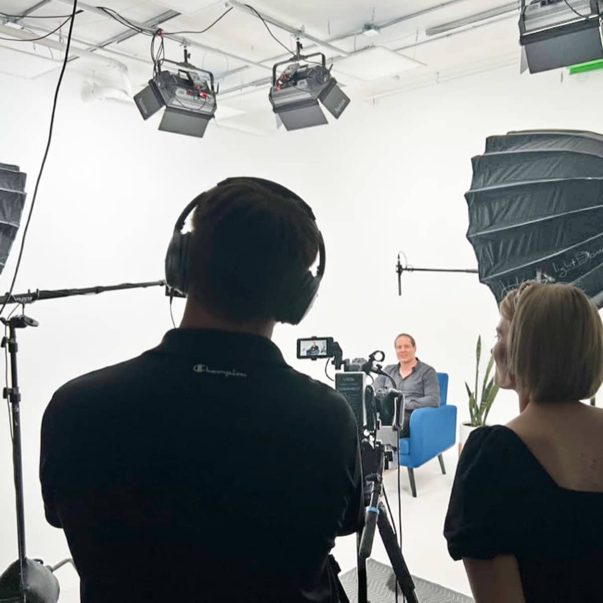 A video recording setup shows two crew members operating a camera and sound equipment, while a seated person is visible in the background.