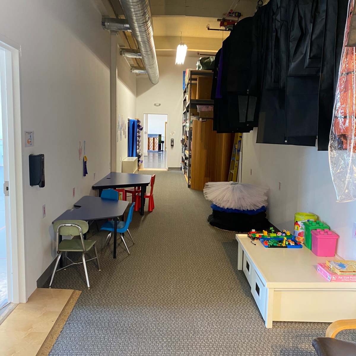 A long, narrow room with a carpeted floor, lined with storage shelves, a table and chairs on one side, and various toys on a low table.