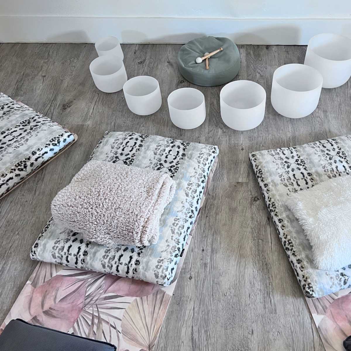 A serene wellness setup with two padded mats, each topped with soft blankets, flanked by several crystal singing bowls arranged in a semi-circle.