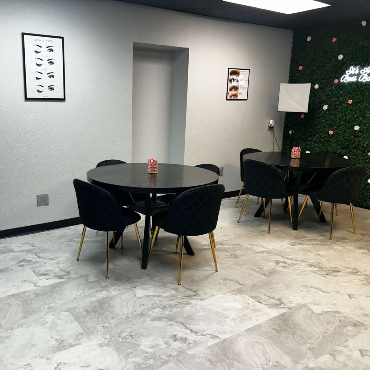 A modern, minimalist waiting area with two circular tables surrounded by black chairs, set against a light gray wall and a greenery accent wall.
