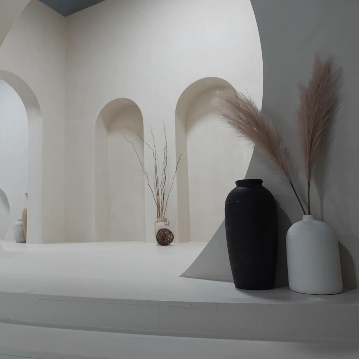 A minimalist interior space with arches, white walls, and various decorative vases and dried plants.