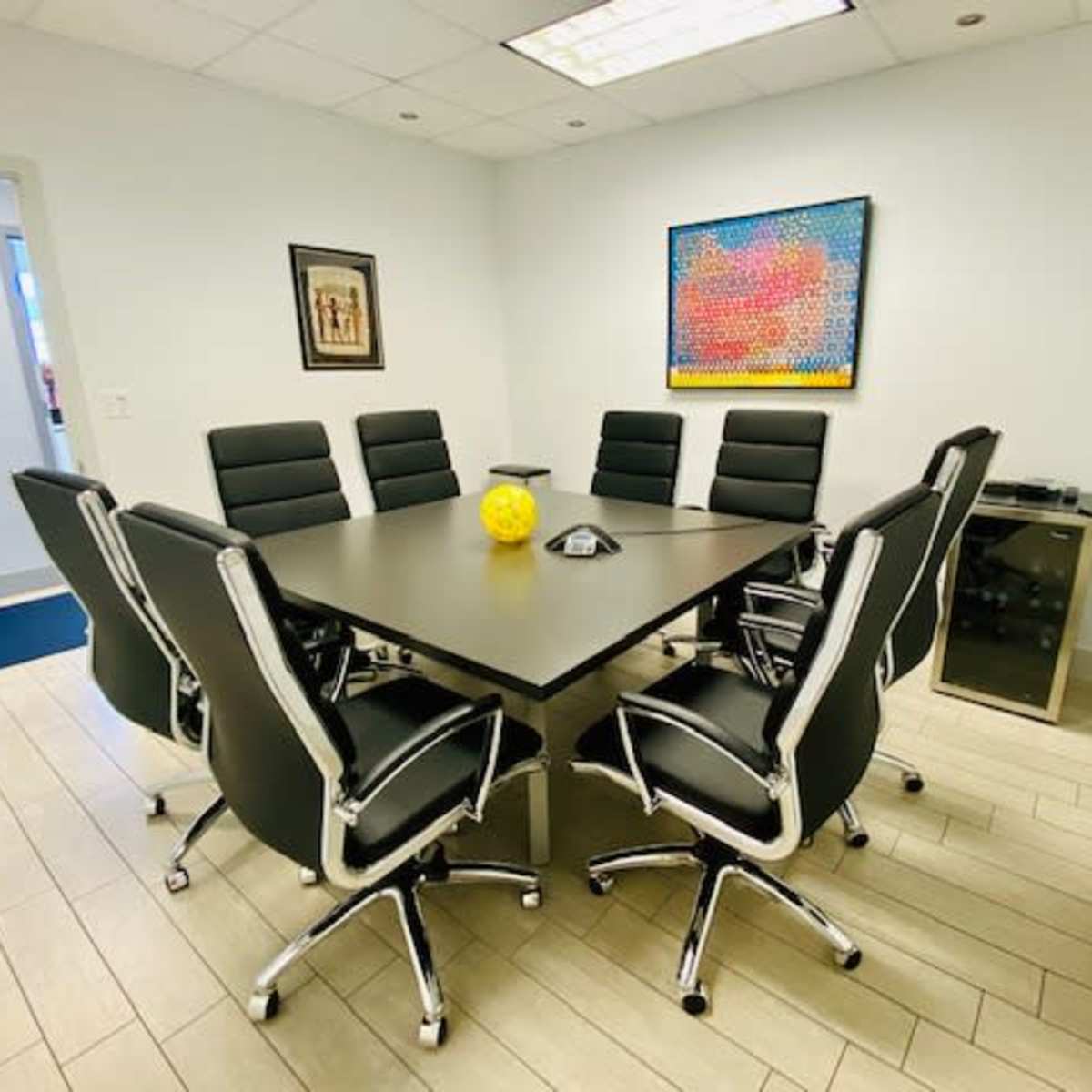 A conference room features a large black table surrounded by eight black office chairs, with a yellow decorative object in the center and colorful artwork on the wall.