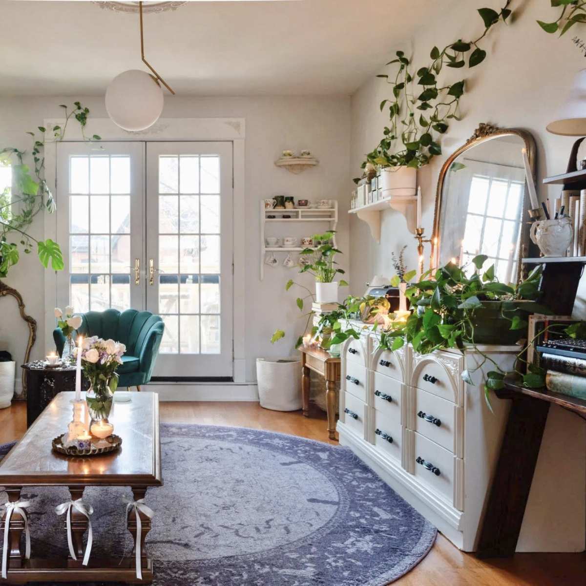 A cozy living room with a vintage sofa, a round coffee table, and several houseplants adorning the shelves and walls, along with large double doors leading to a balcony.