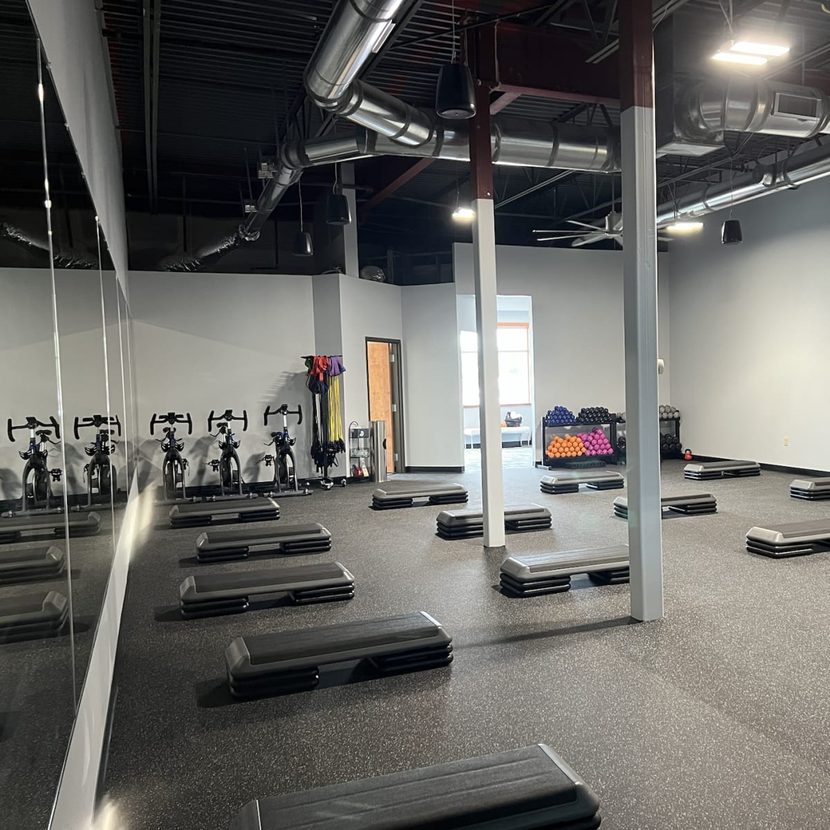 A spacious fitness studio with numerous step platforms arranged on the black rubber floor, exercise bikes lined up against the wall, and a few weights scattered in one corner.