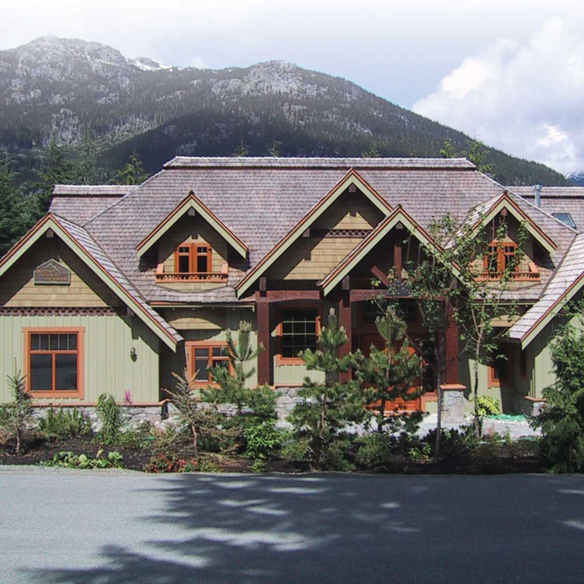 A large, multi-roofed house featuring a combination of green and tan siding, set against a backdrop of mountains and trees.