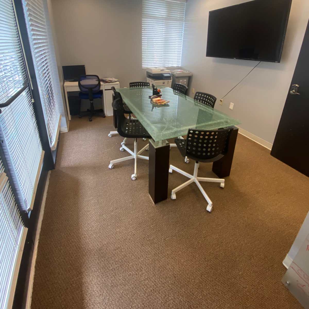 A conference room featuring a glass table surrounded by black rolling chairs, a large wall-mounted screen, and a window with blinds.