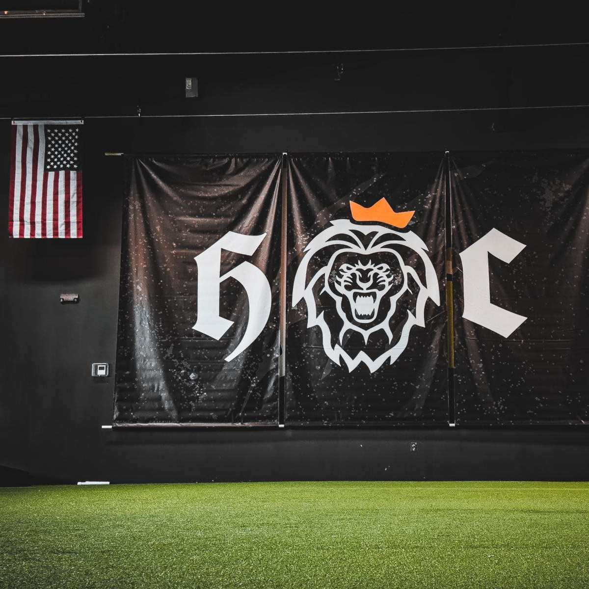 A large black banner with a lion's head logo and the letters "H" and "C" in a gym setting, flanked by an American flag on the left and a banner reading "JESUS IS KING" on the right.