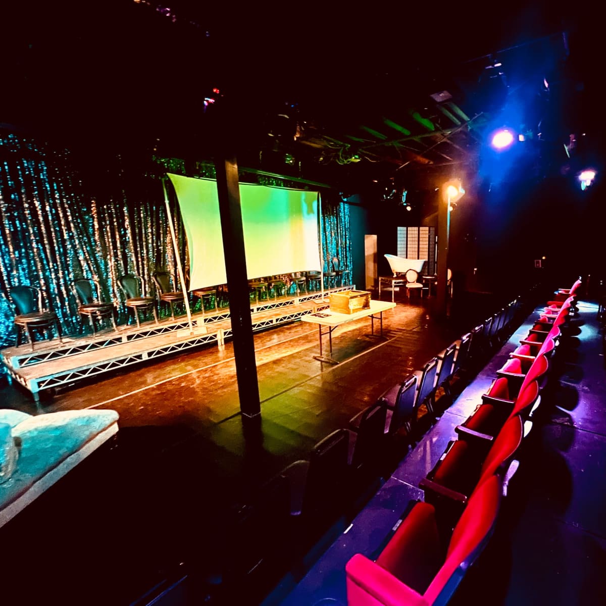 A dimly lit theater with red seating facing a stage that has a large screen and a backdrop adorned with shimmering curtains.