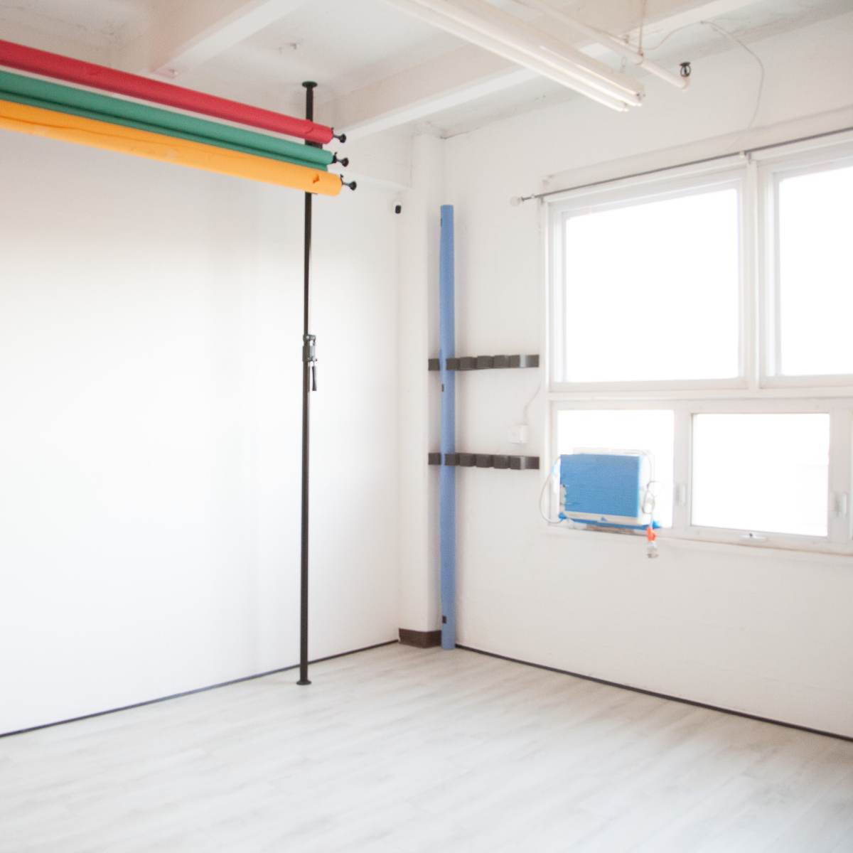 A bright, empty studio space with a blank white wall and colorful backdrops hanging on a vertical support.