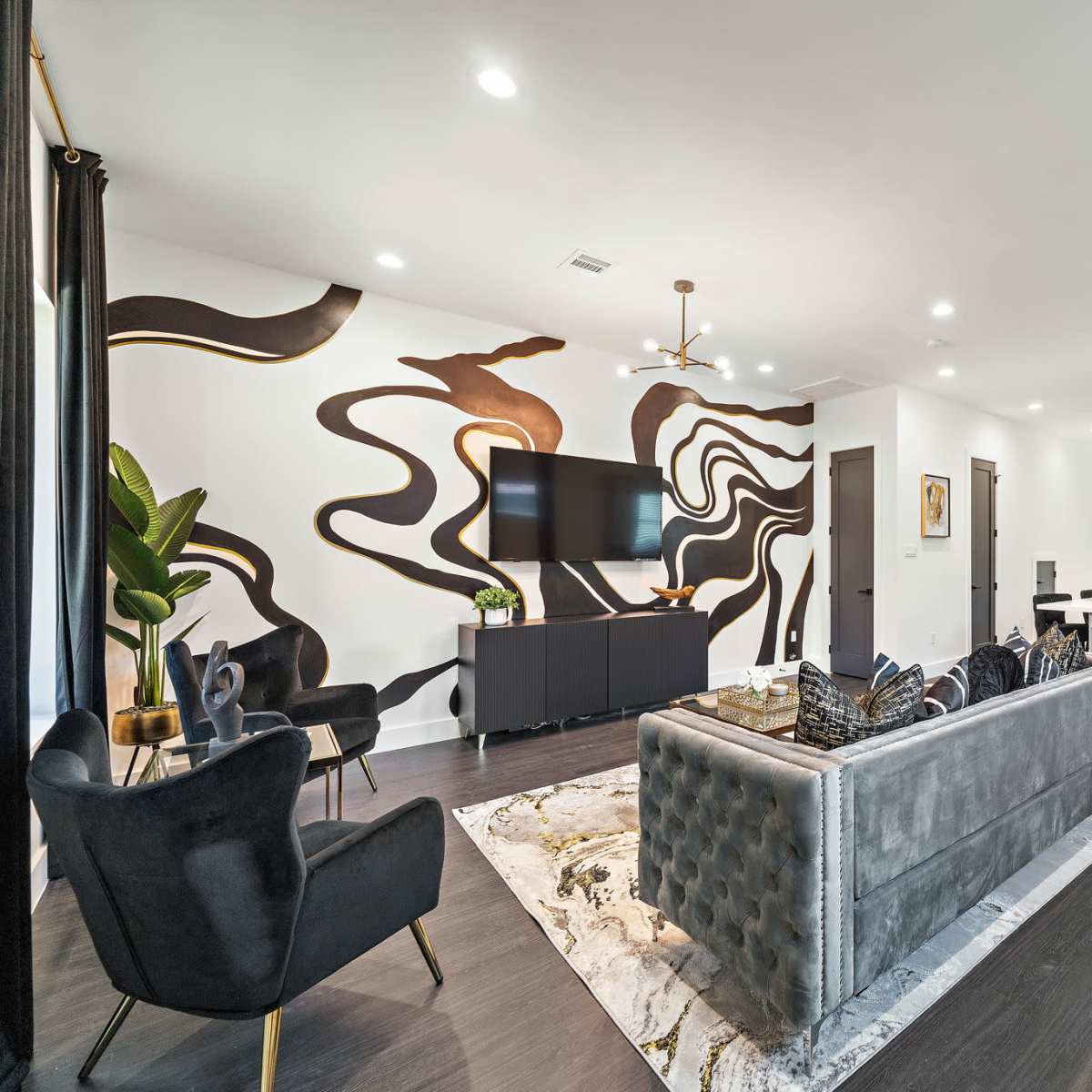 A modern living room with a sectional sofa, a large wall-mounted TV, and a decorative mural on one wall, leading into a kitchen area.