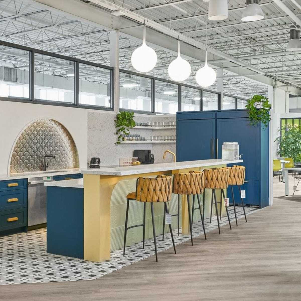 A modern kitchen area with a blue island, gold accents, and bar stools, surrounded by large windows and greenery.