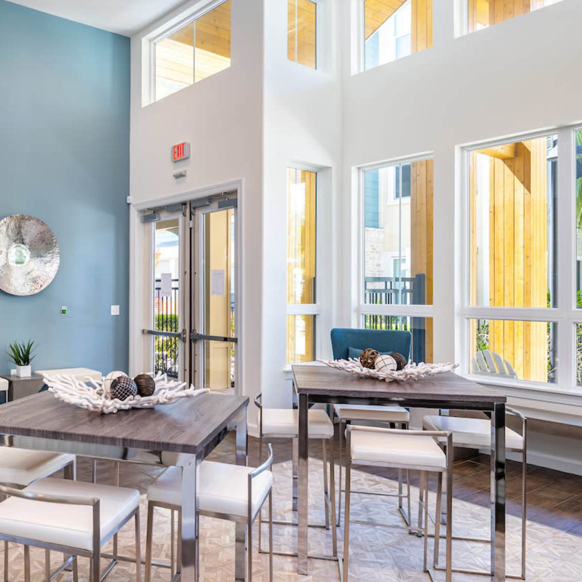 A modern lobby area with tables and chairs, large windows allowing natural light in, and a decorative wall piece.