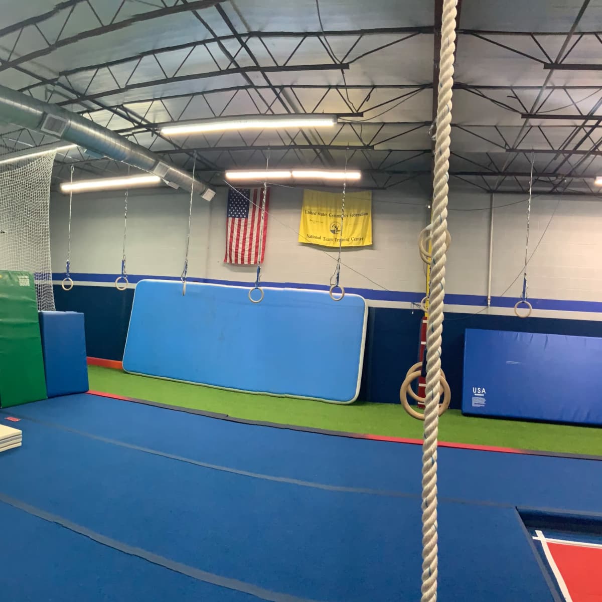 An indoor gymnasium equipped with gymnastic mats, ropes, and various training apparatuses along with basketball hoops and a safety netting system.