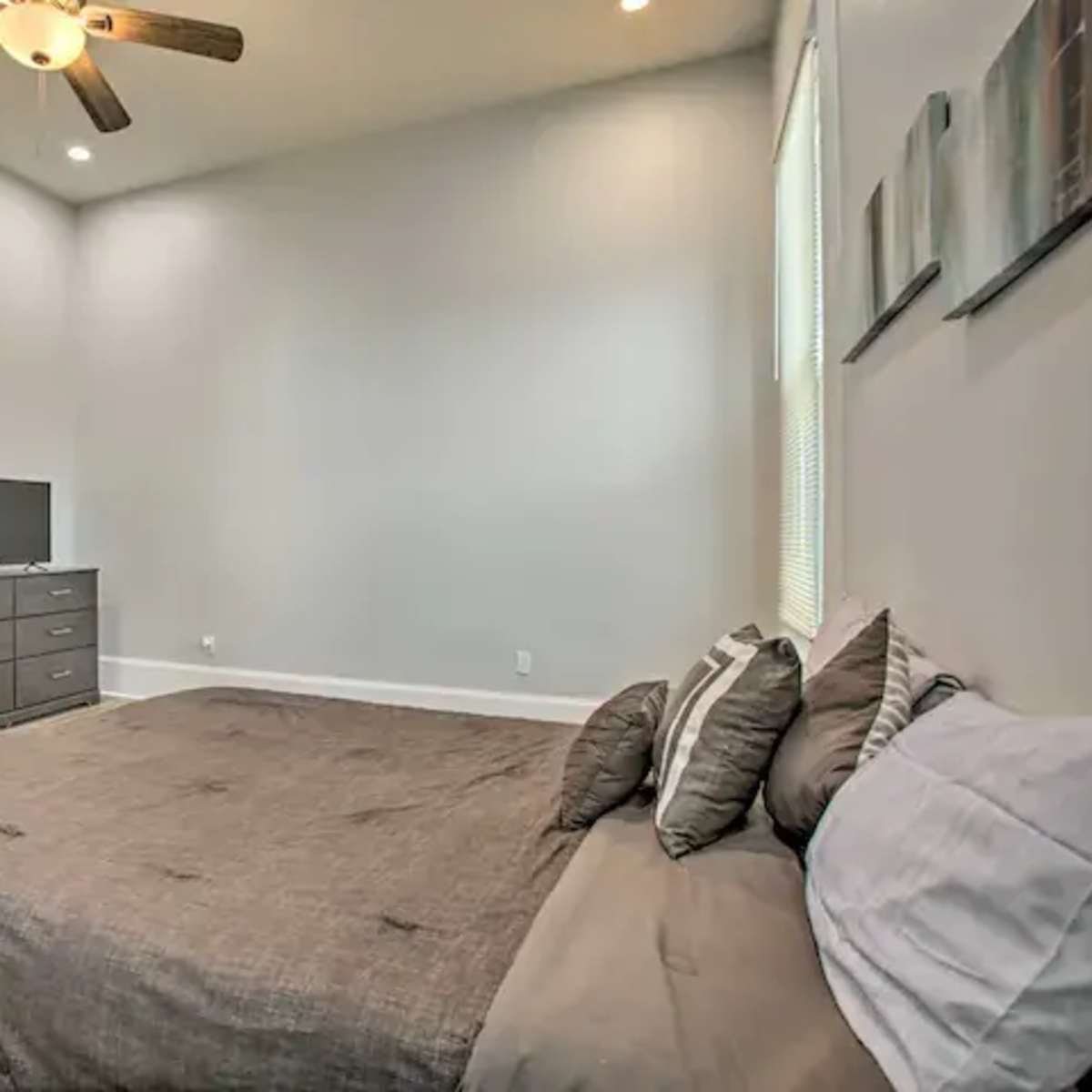 A tidy bedroom with a gray bedspread, a dresser, a television, and natural light coming through the windows.