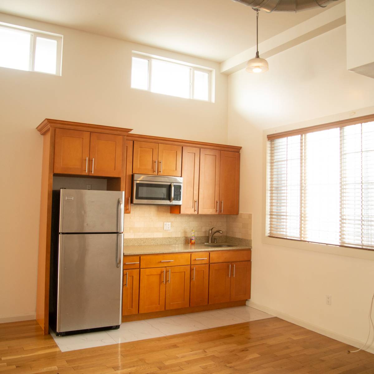A modern kitchen featuring wooden cabinets, a stainless steel refrigerator, and a countertop with a sink, all illuminated by natural light from the windows.