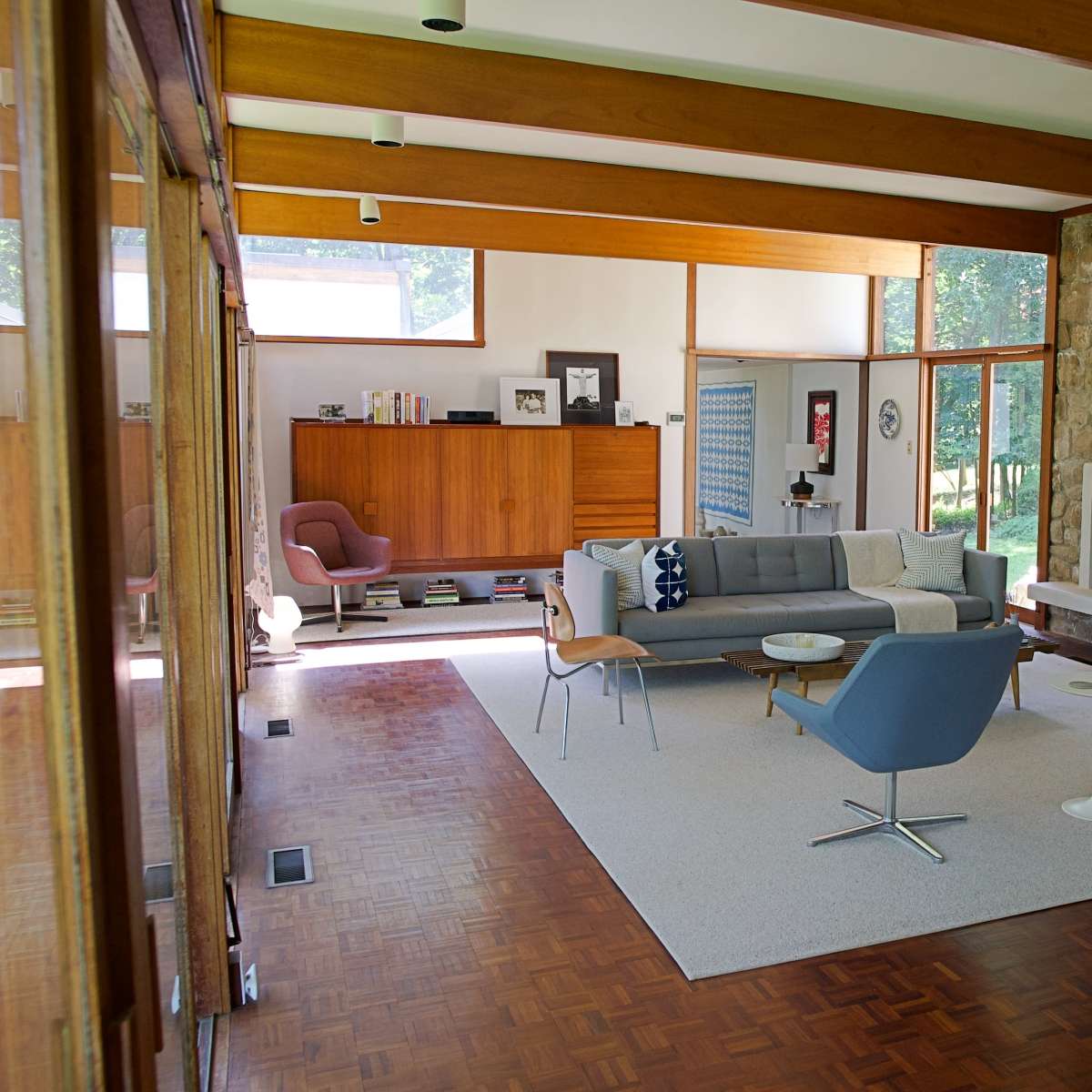A modern living room with a stone fireplace, a gray sofa, two blue chairs, and large windows allowing natural light to enter.