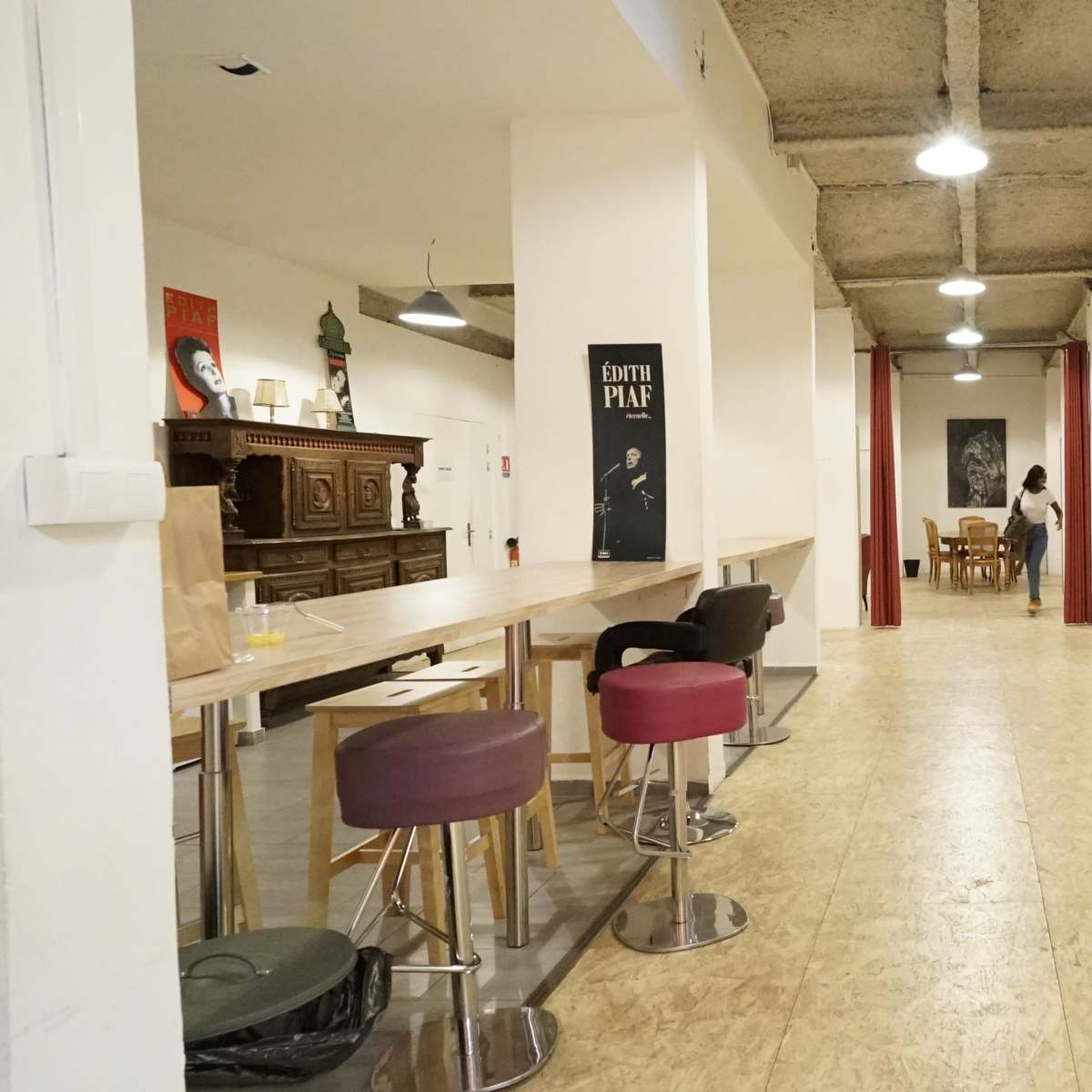 A spacious interior with a long table and stools on the left, wooden tables and chairs scattered throughout, and white walls with artwork displayed.