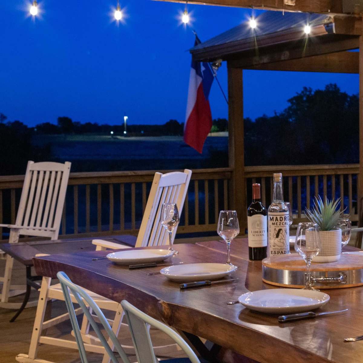 A wooden dining table is set with plates and glasses on a porch at dusk, featuring a Texas flag in the background.