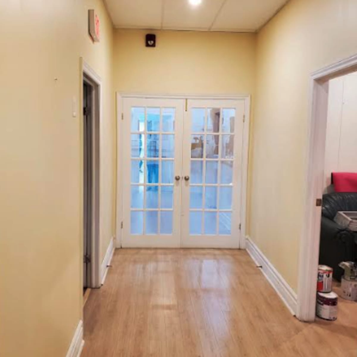 A long, narrow hallway with yellow walls leads to a set of French doors at the end, with a view of a room beyond.