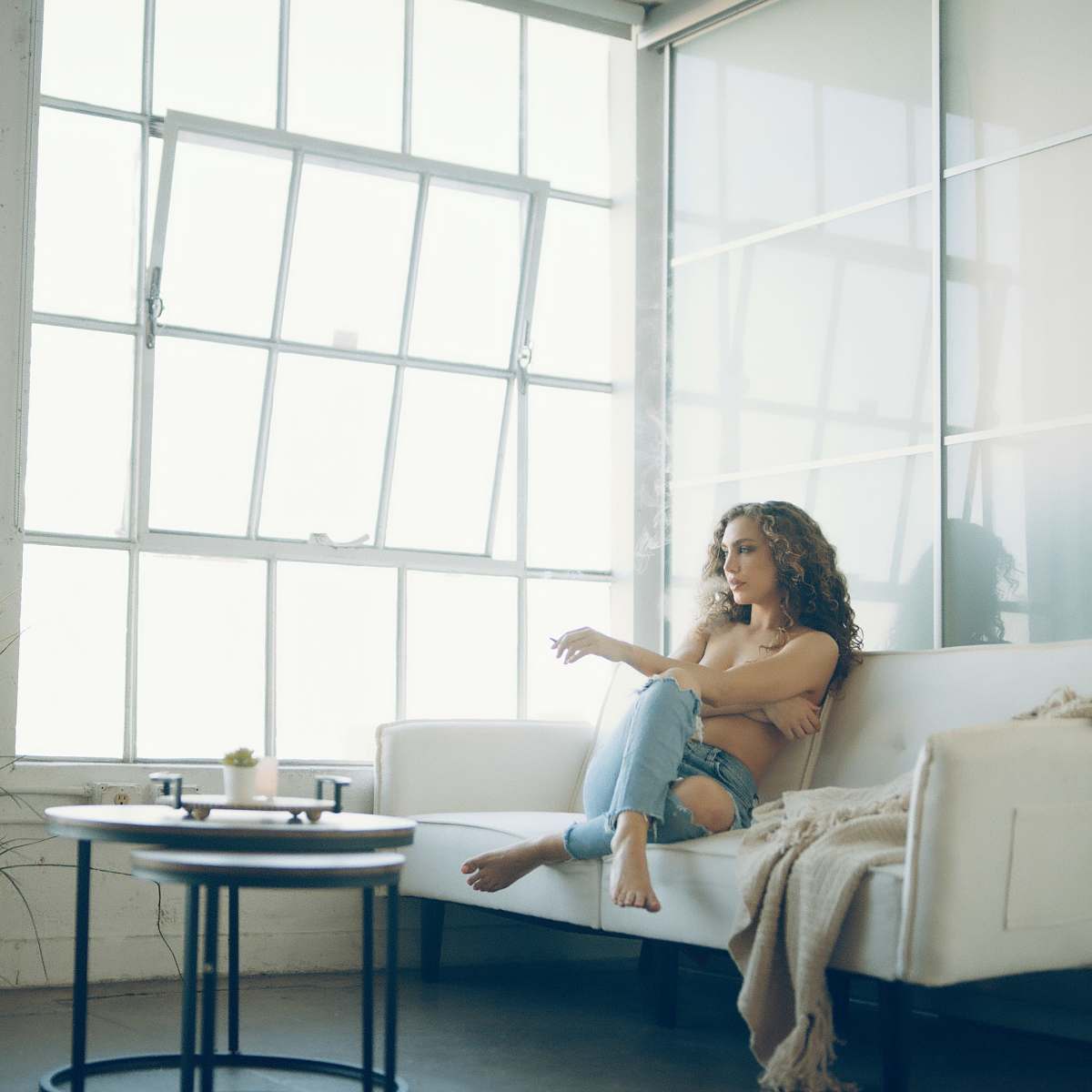 A woman sits on a white couch in a bright room with large windows, facing a small coffee table.