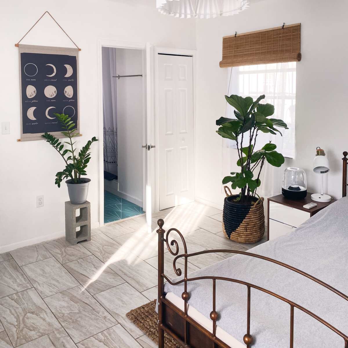 The room features a bed with a metal frame, a large pendant light, potted plants in the corners, and a door leading to a bathroom.