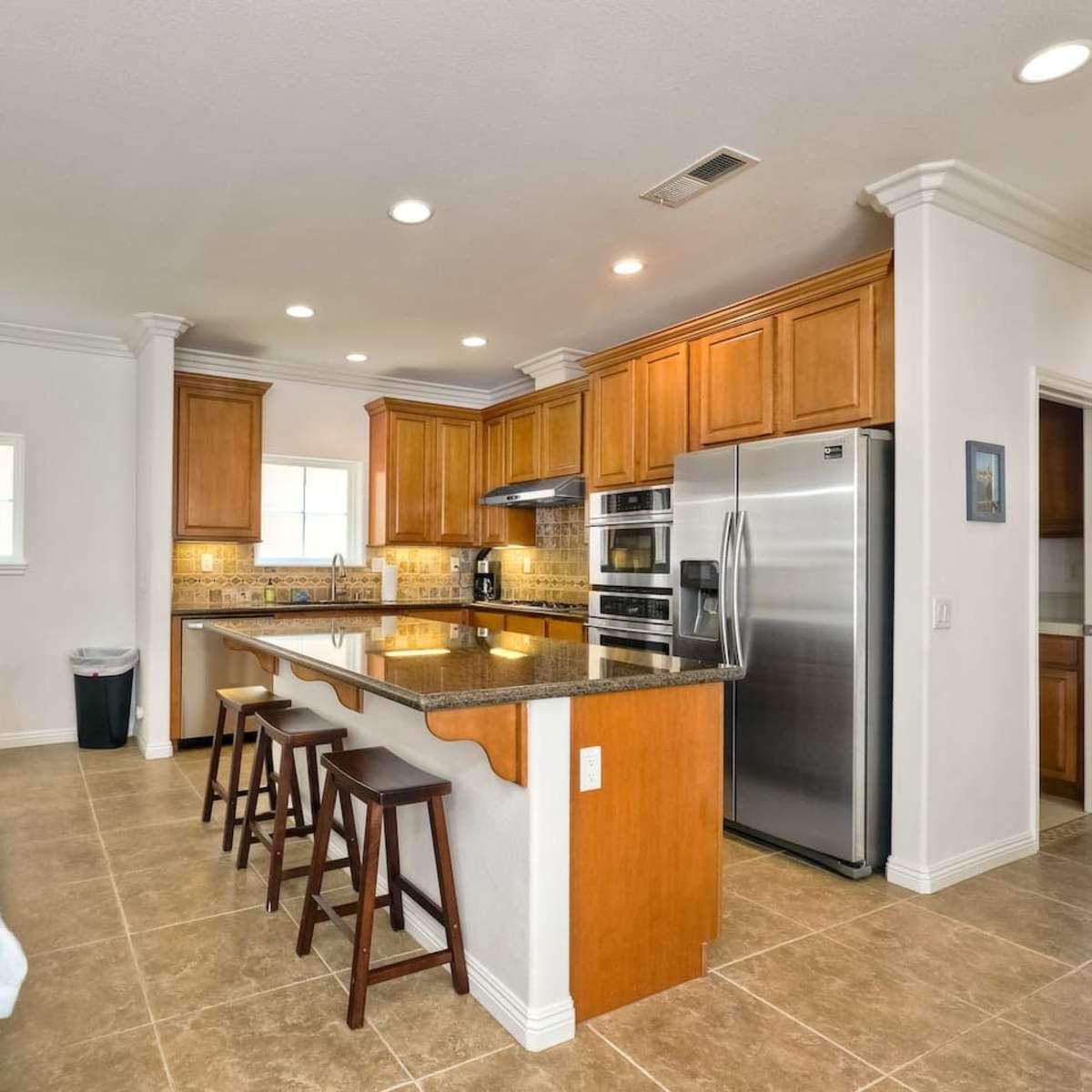 A modern kitchen with wooden cabinets, stainless steel appliances, and a central island with bar stools, adjacent to a living area with a gray sofa.