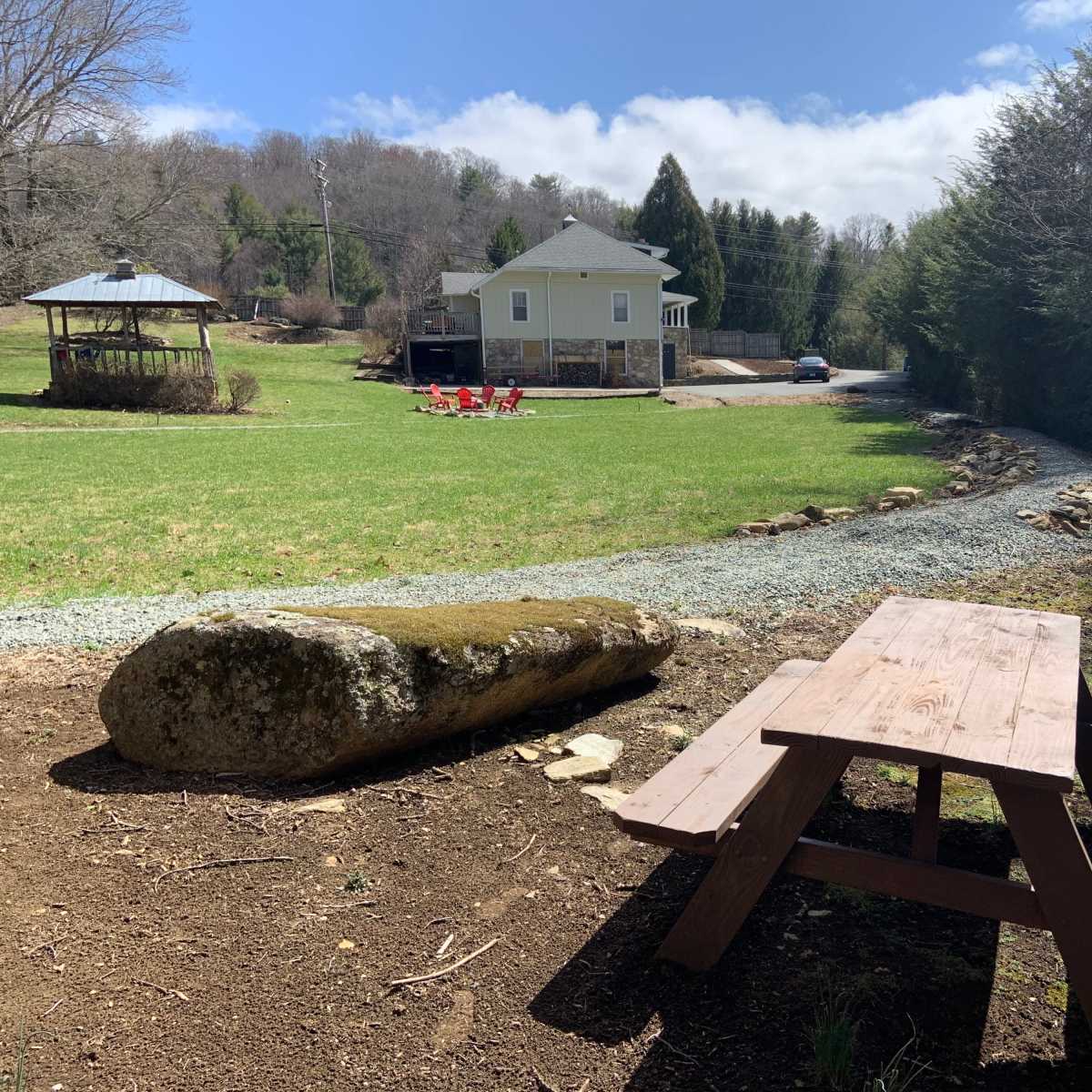 A wooden picnic table sits on a dirt area near a grassy lawn, with a stone and gravel path leading to a house and gazebo in the background.