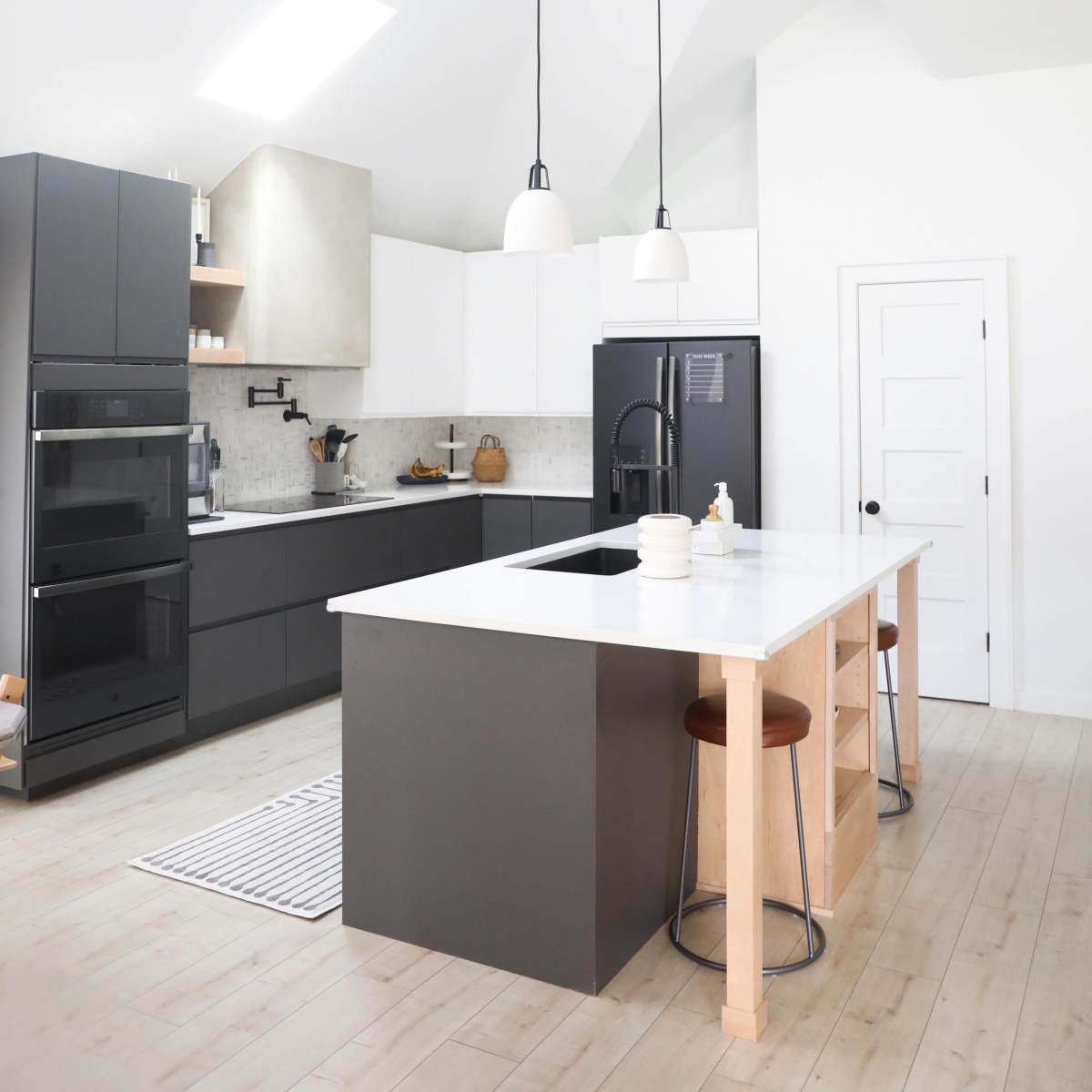 A modern kitchen featuring a central island with two stools, black cabinets, light countertops, and skylights above.