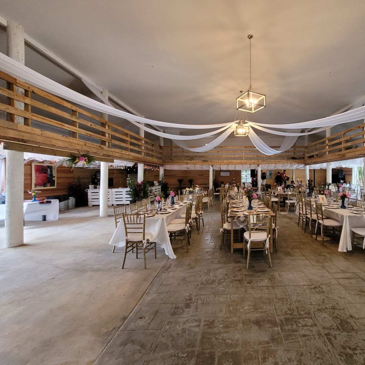 A spacious indoor event venue with neatly arranged tables covered in white linens and decorations, featuring a rustic wooden decor and a high ceiling with draped fabric.