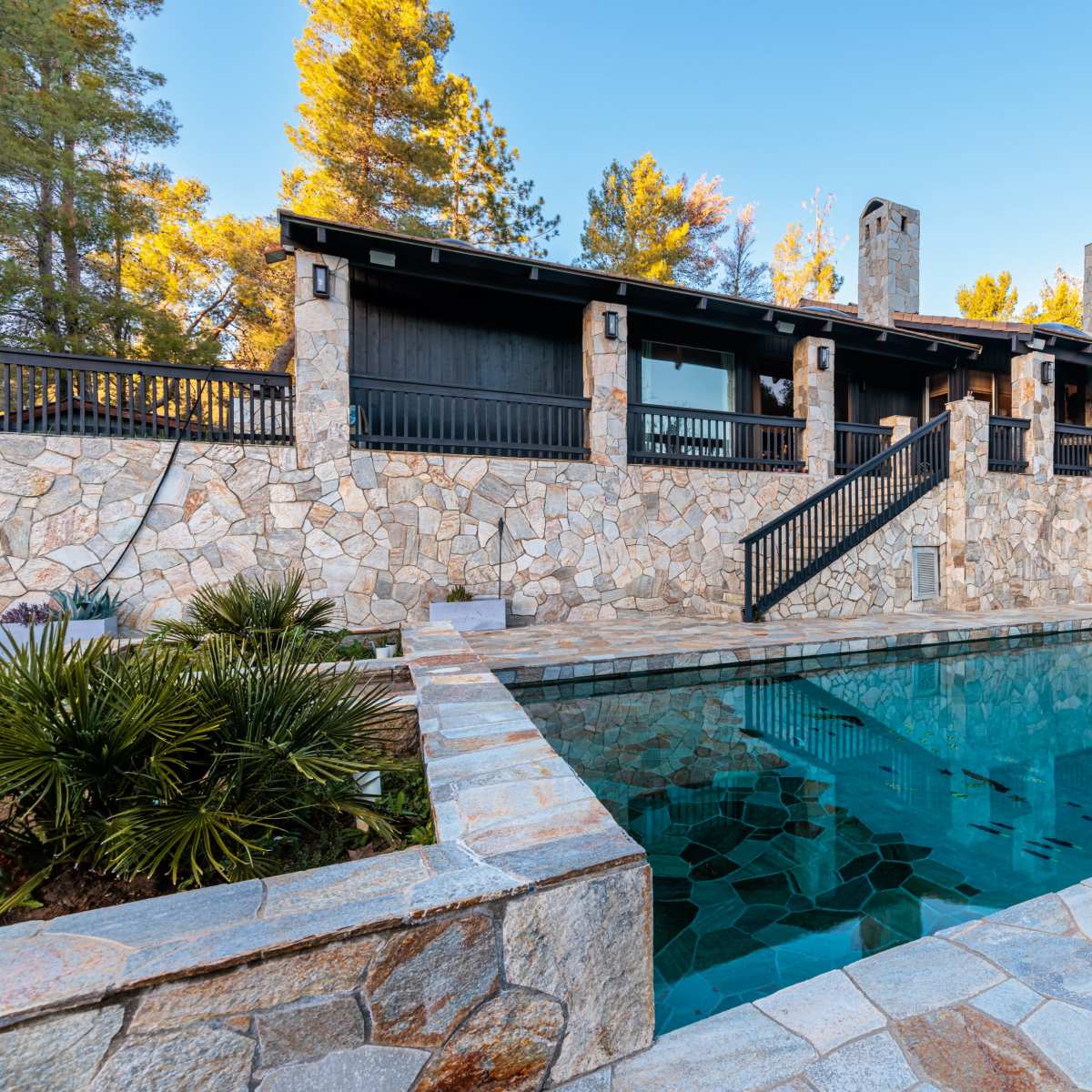 A stone-lined swimming pool in front of a rustic, two-story house surrounded by trees.